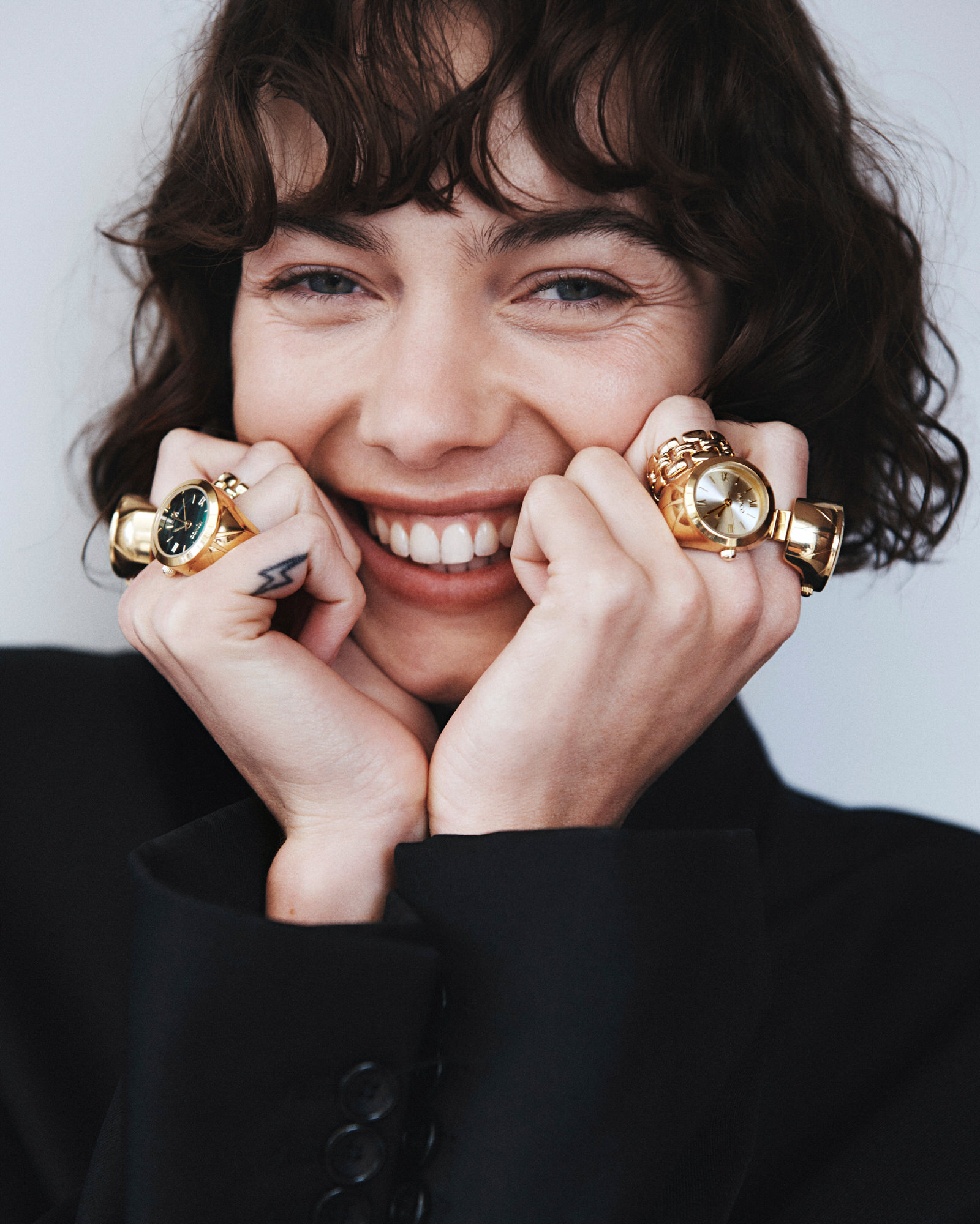 Smiling woman with dark curly hair wearing gold watch rings and a black blazer against a plain white background.