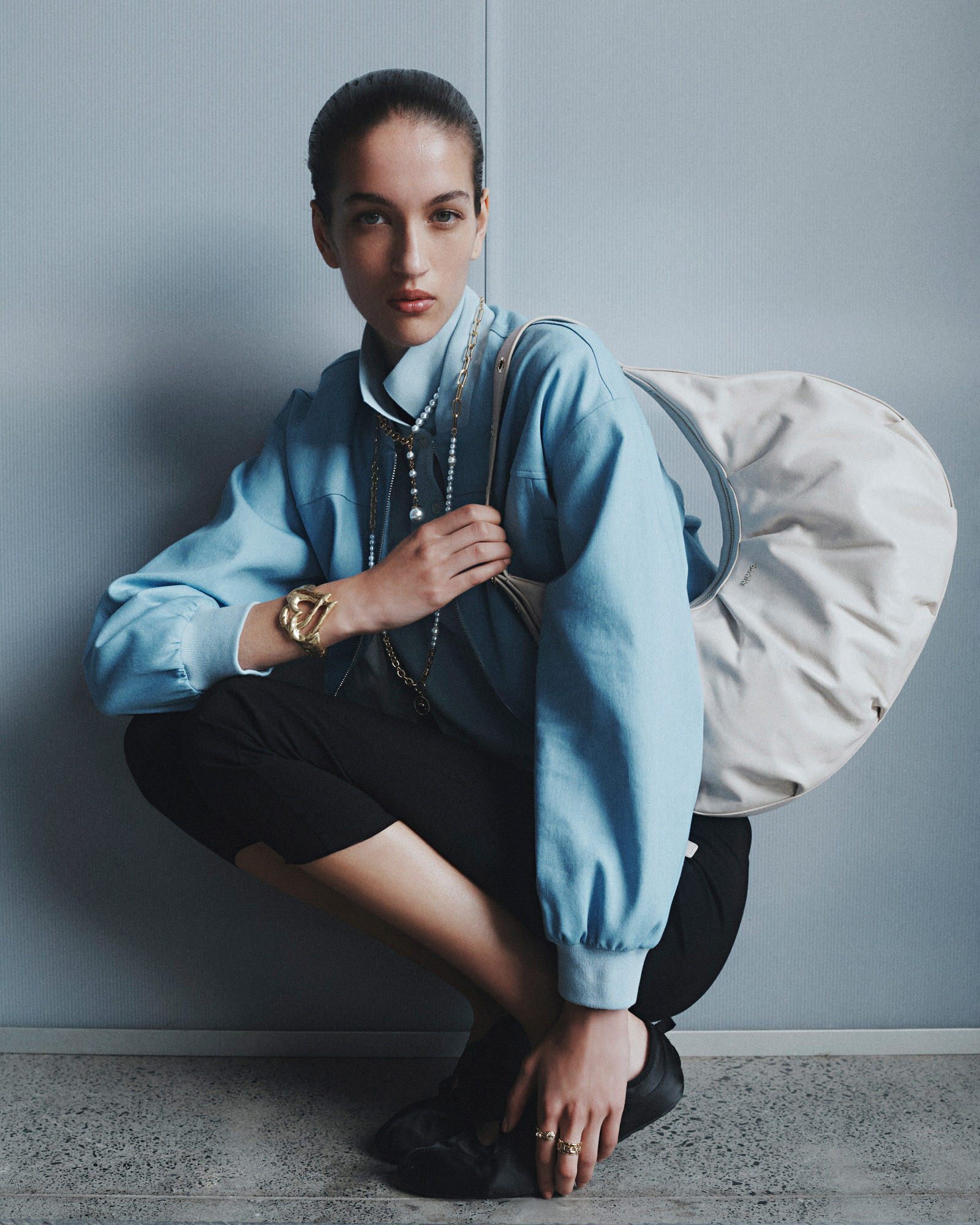 A young woman with styled hair poses in a photo studio, wearing a pale blue long-sleeved blouse, dark skirt, gold jewelry, and carrying a white bag.