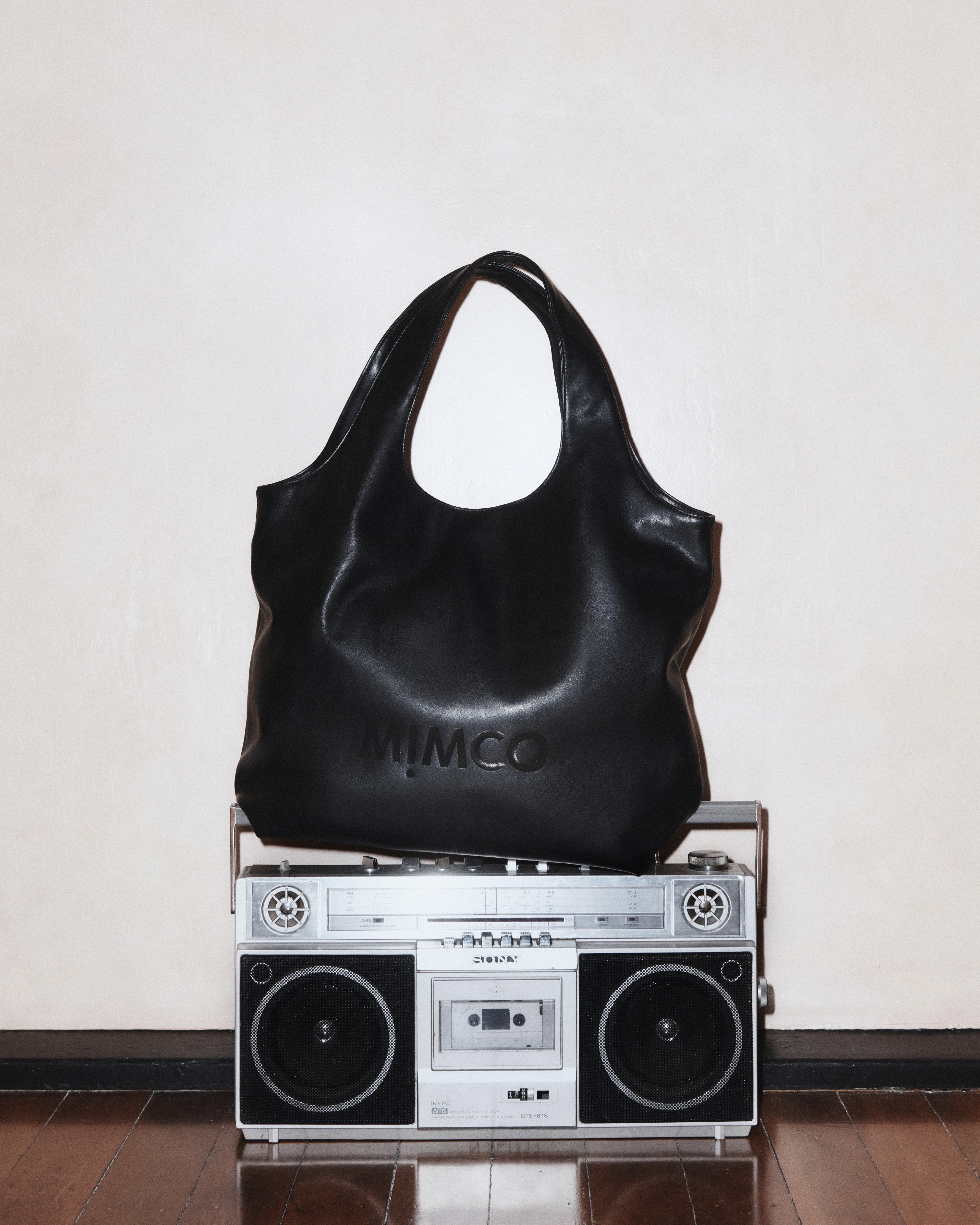 Black leather Mimco handbag sits atop a vintage boombox against a neutral wall, showcasing fashion and retro technology.