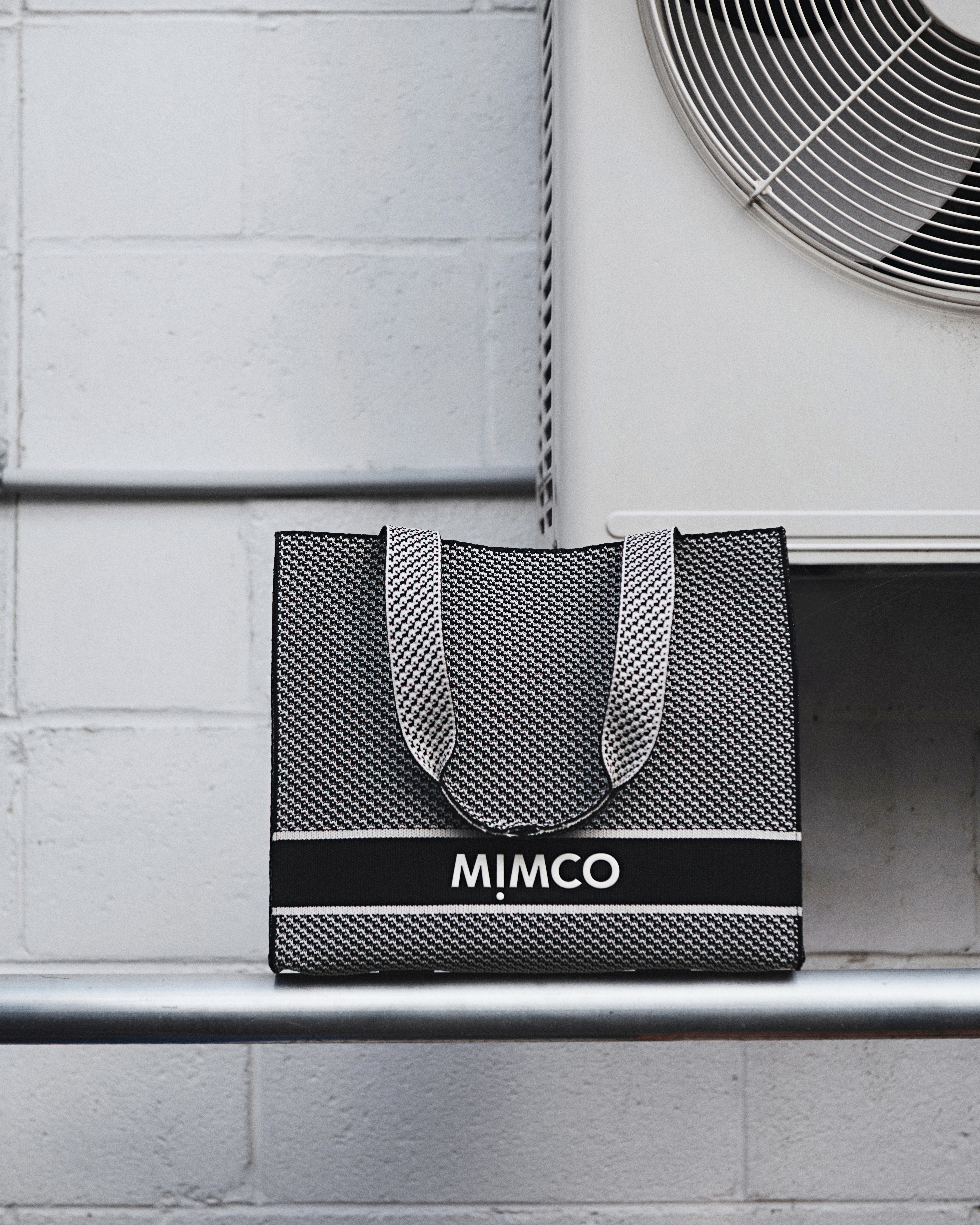 Black and white woven Mimco tote bag resting on a horizontal metal pipe against a white block wall next to an air conditioning unit.