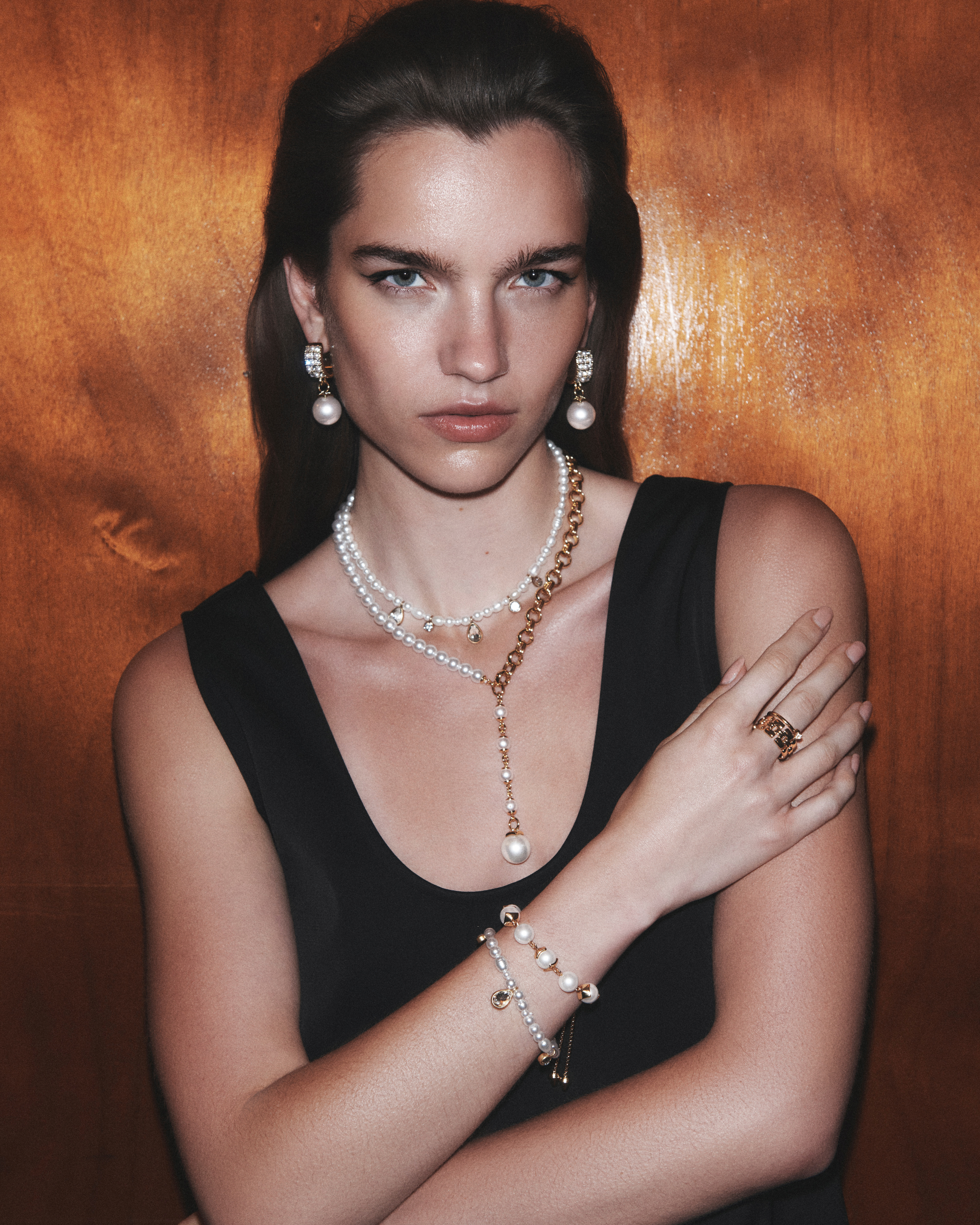 Portrait of a woman in a black dress adorned with pearl jewelry, including earrings, necklace, and bracelet, against a bronze backdrop.