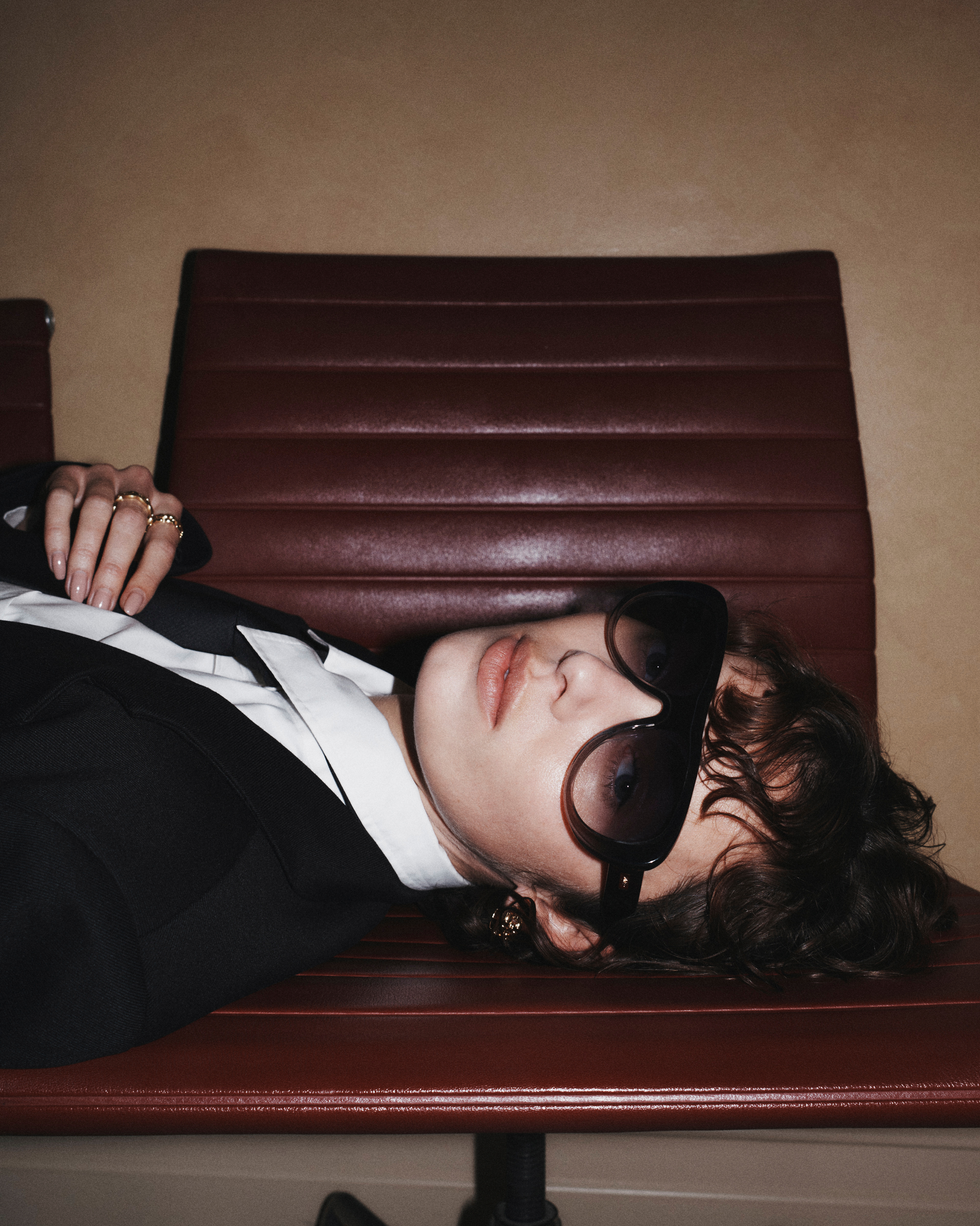 Stylish young woman in sunglasses and a suit jacket lounges on a burgundy leather bench against a neutral-toned wall in a studio setting.