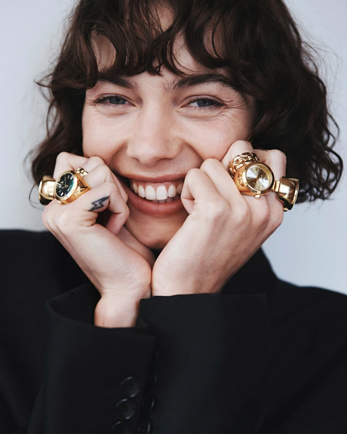Smiling woman with dark curly hair wearing gold watch rings and a black blazer against a plain white background.