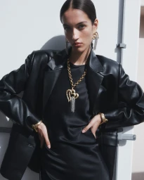 Woman in an oversized black leather blazer and black dress wearing a large gold heart statement necklace and matching drop earrings.