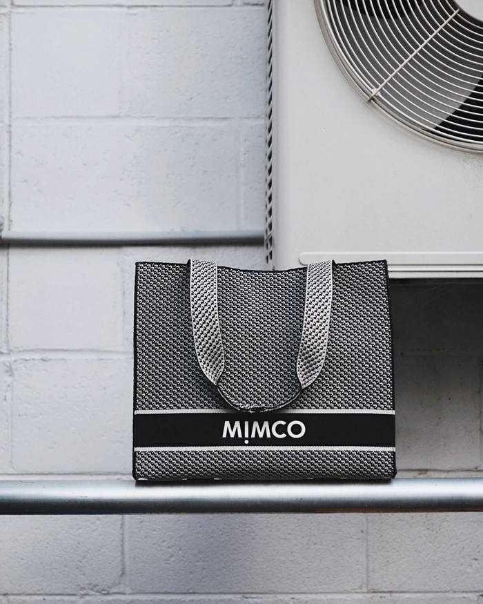 Black and white woven Mimco tote bag resting on a horizontal metal pipe against a white block wall next to an air conditioning unit.