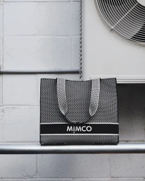 Black and white woven Mimco tote bag resting on a horizontal metal pipe against a white block wall next to an air conditioning unit.