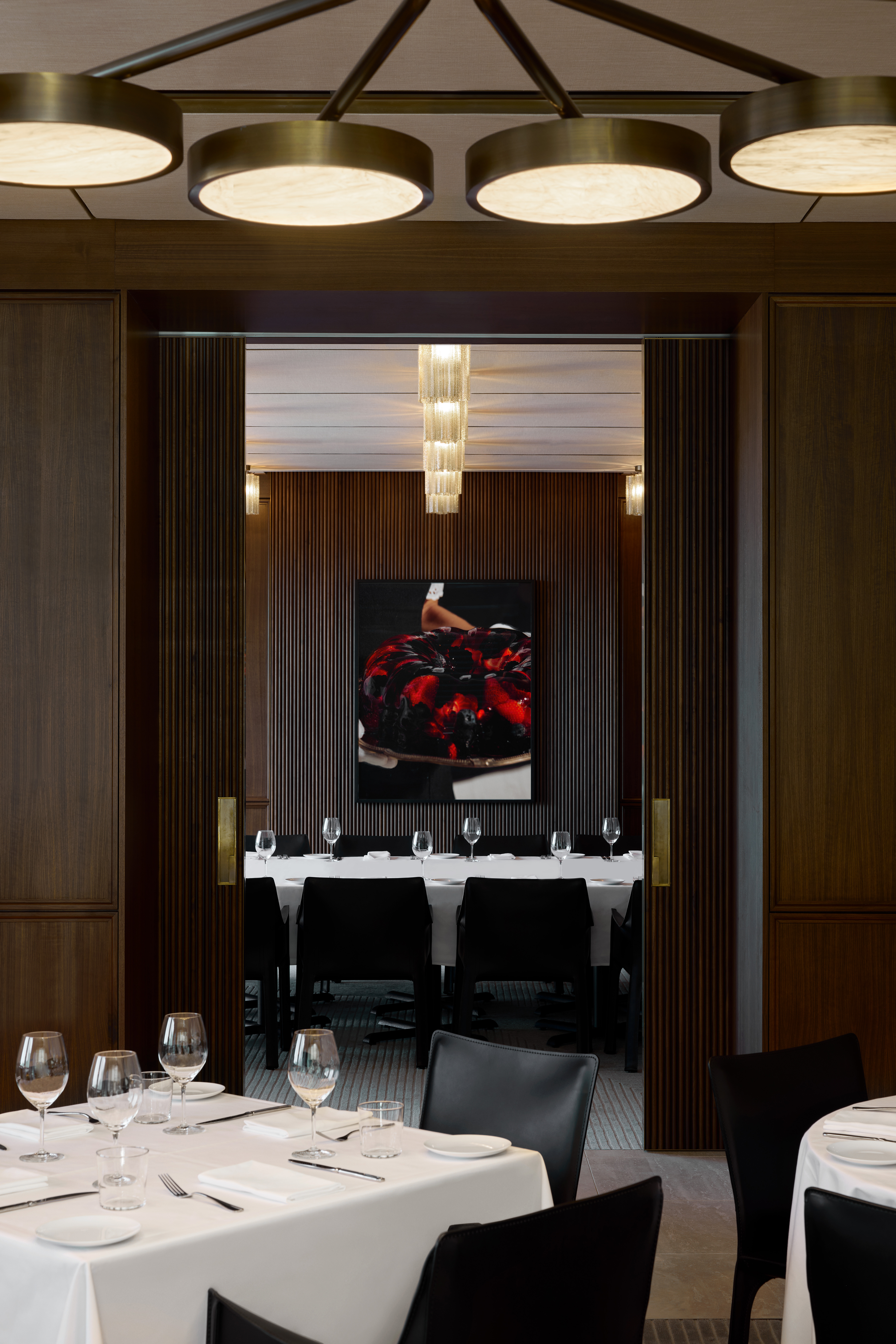 Interior view of a fine dining restaurant featuring set tables with wine glasses, modern lighting, and a painting viewed through doorway.