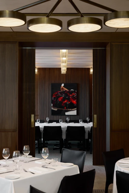 Interior view of a fine dining restaurant featuring set tables with wine glasses, modern lighting, and a painting viewed through doorway.