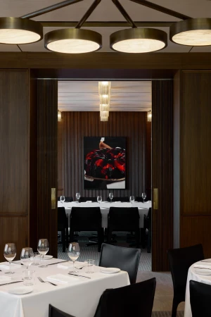 Interior view of a fine dining restaurant featuring set tables with wine glasses, modern lighting, and a painting viewed through doorway.