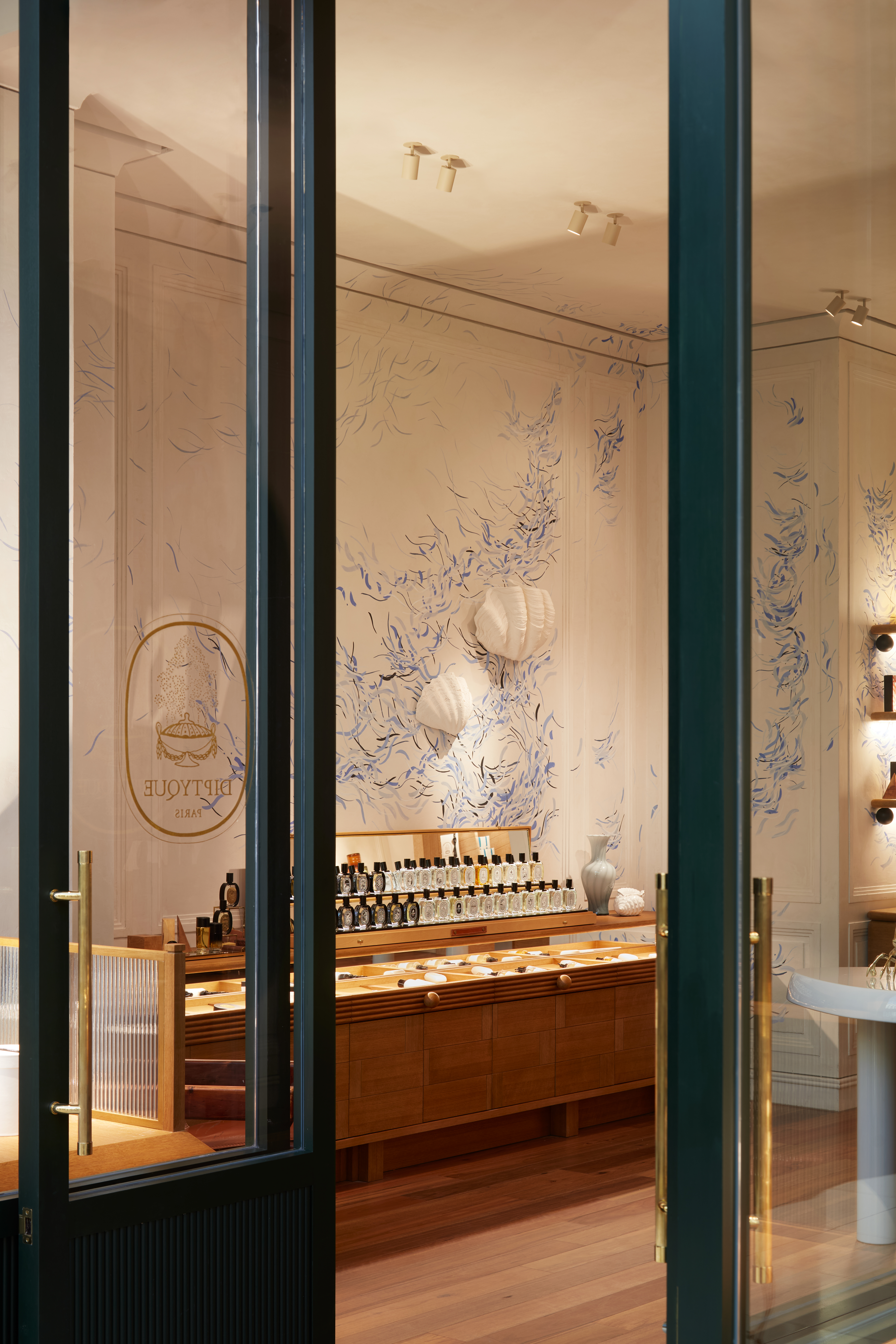 Interior of a perfume shop with shelves and display case, featuring a white vase and botanical wall art, viewed through a glass door.