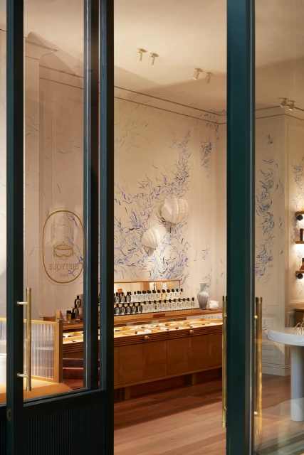 Interior of a perfume shop with shelves and display case, featuring a white vase and botanical wall art, viewed through a glass door.