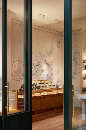 Interior of a perfume shop with shelves and display case, featuring a white vase and botanical wall art, viewed through a glass door.