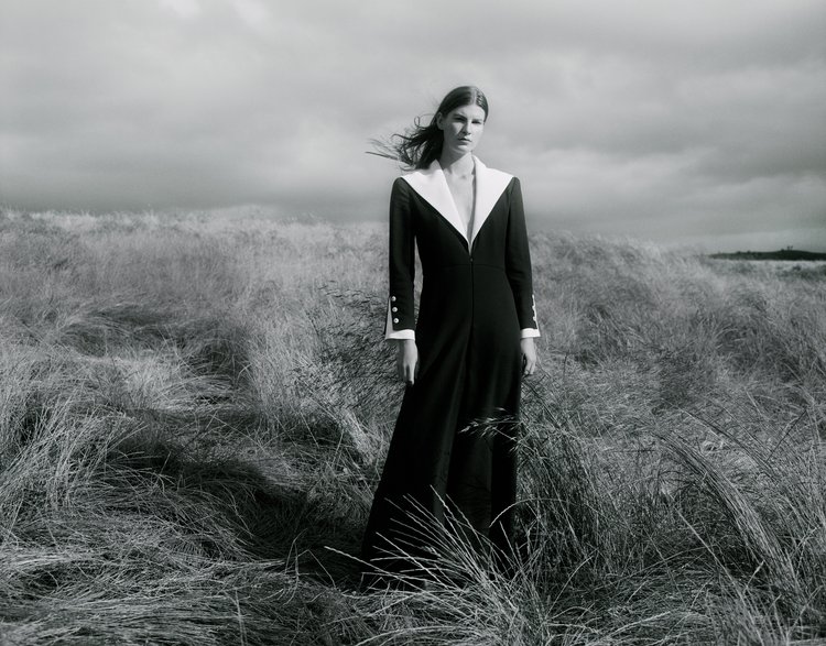 A woman in a long black dress stands in a field of tall, windblown grasses, against a grey sky. The image is black and white.