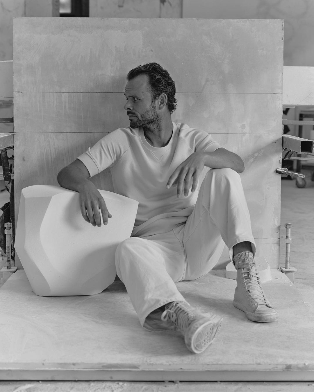 Man sitting next to a geometric object in a monochromatic indoor scene, wearing casual clothing and sneakers, set against a neutral backdrop.