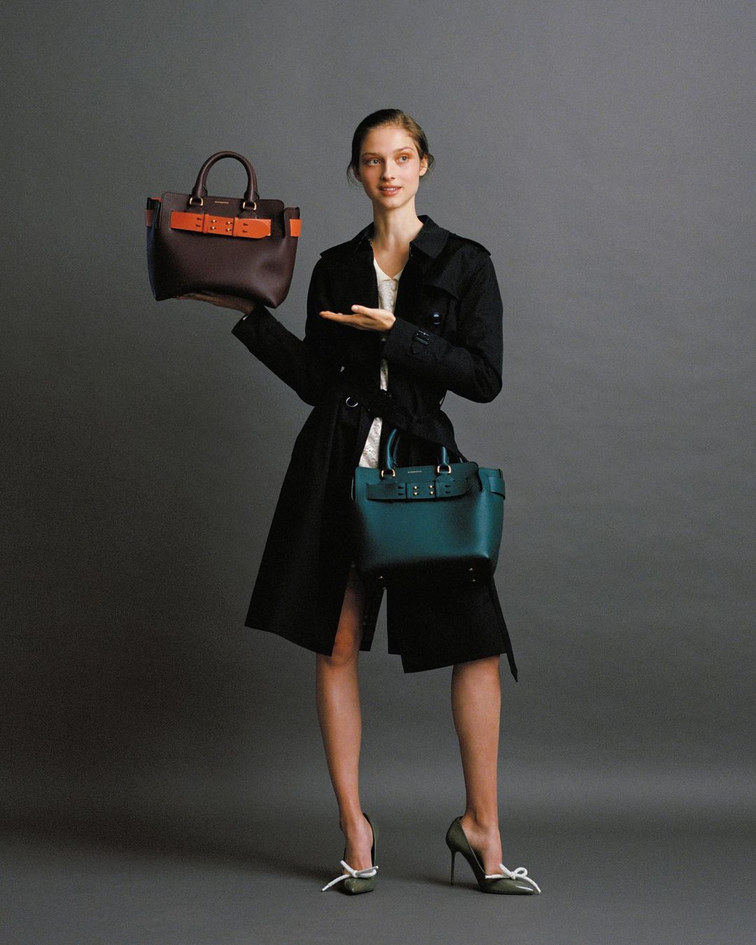 Adult female fashion model poses in a studio with a brown handbag in one hand and an aquamarine handbag below.