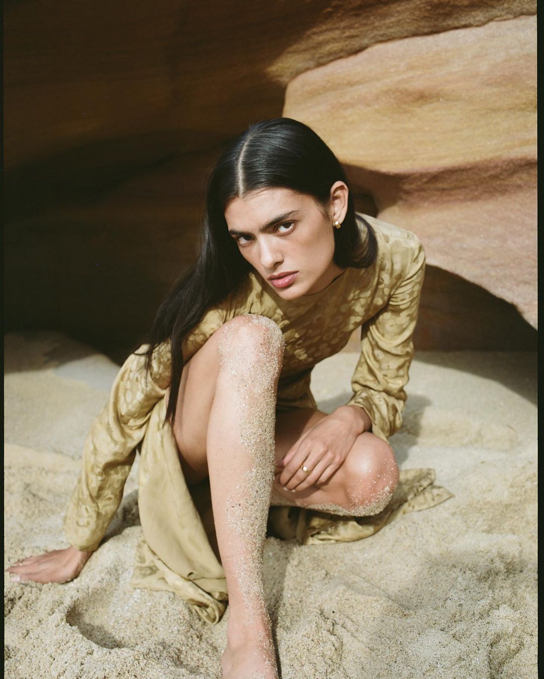 Brunette woman in a golden dress poses in sand against a rock formation, looking at the camera with a neutral expression.