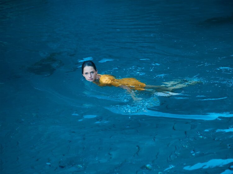 Young woman with wet hair, wearing an orange dress, swims in a blue pool of water, creating ripples and motion in the water.