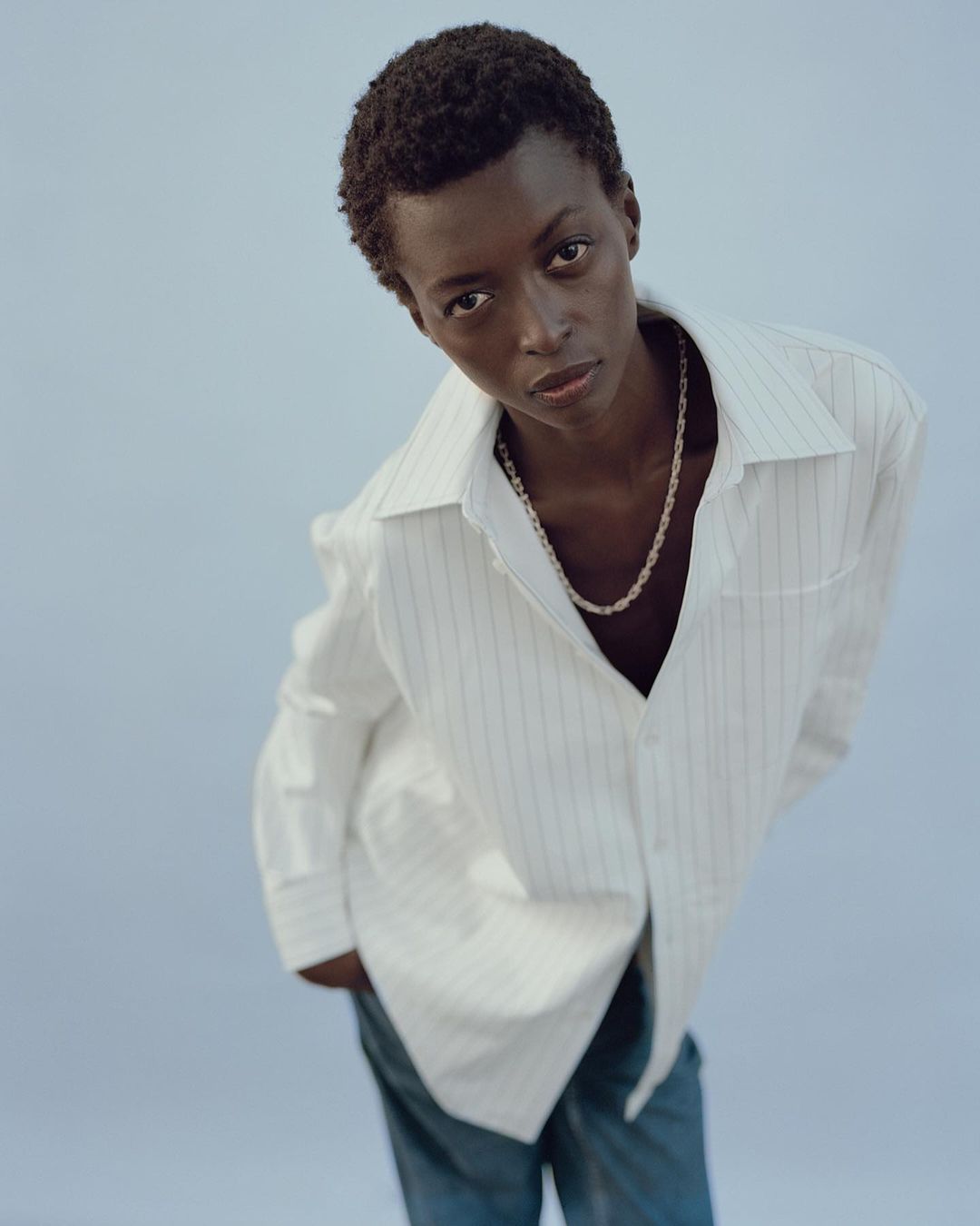 Fashion portrait of a young Black woman in a striped button-down shirt and jeans against a pale blue background, looking at the camera.