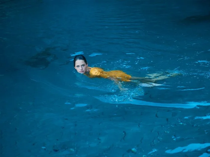 A woman in a yellow dress swimming in deep blue water, styled by Natalie Petrevski.