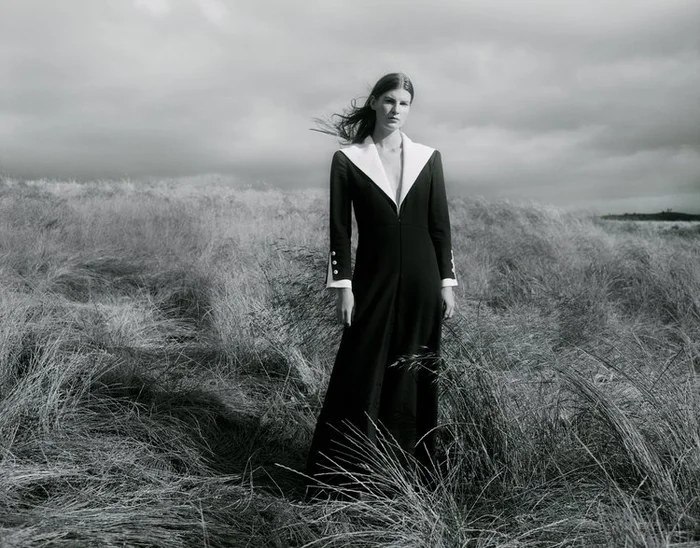 Model wearing a long black coat with white lapels standing in a tall grass field under an overcast sky.