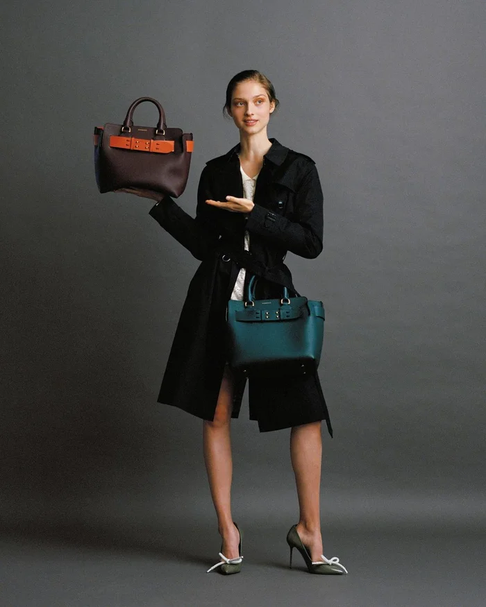 Model wearing a black coat holding two structured leather handbags against a dark grey background by Natalie Petrevski