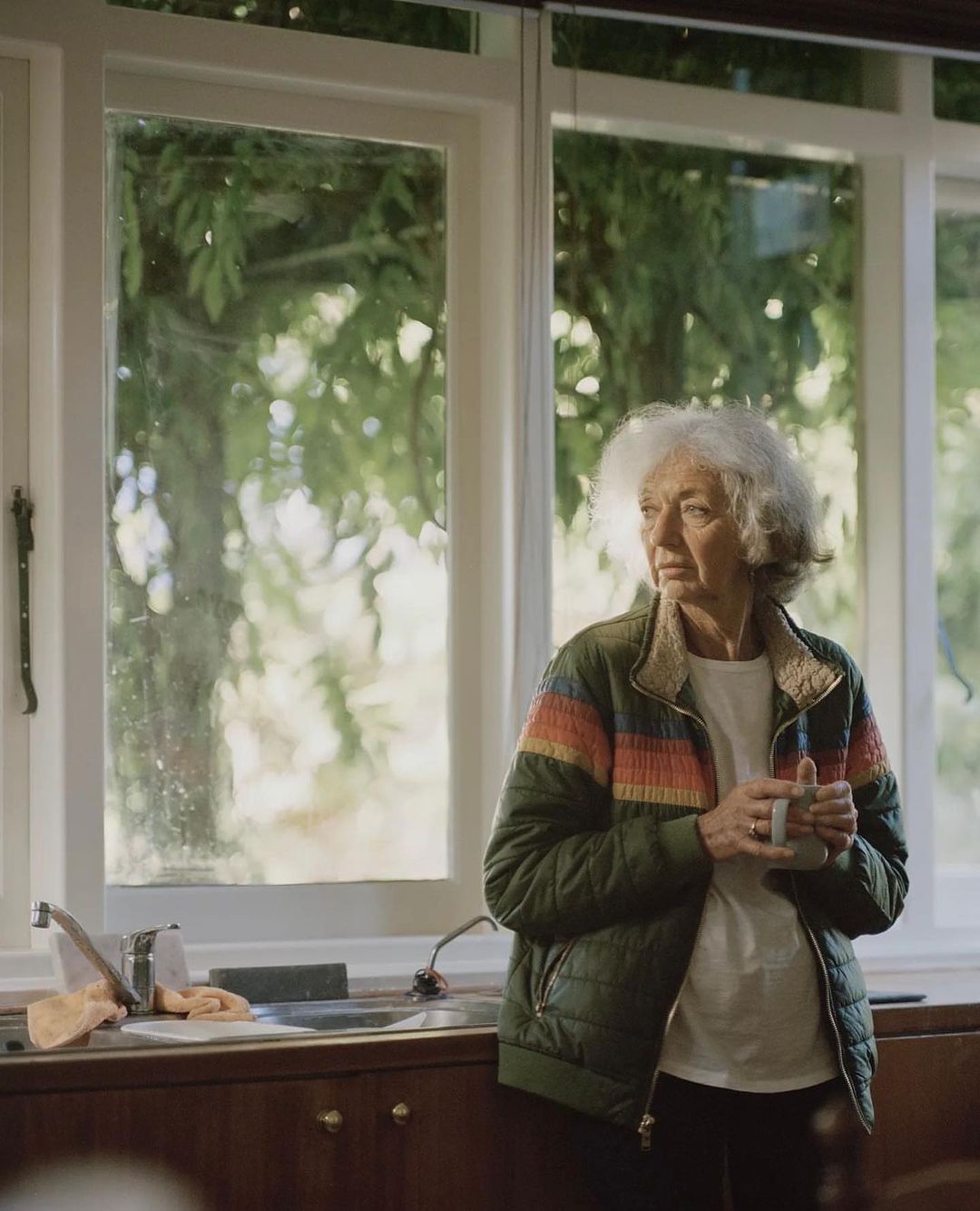 Elderly woman with short white hair stands indoors near a window with a cup in hand, wearing a green quilted jacket in natural light.