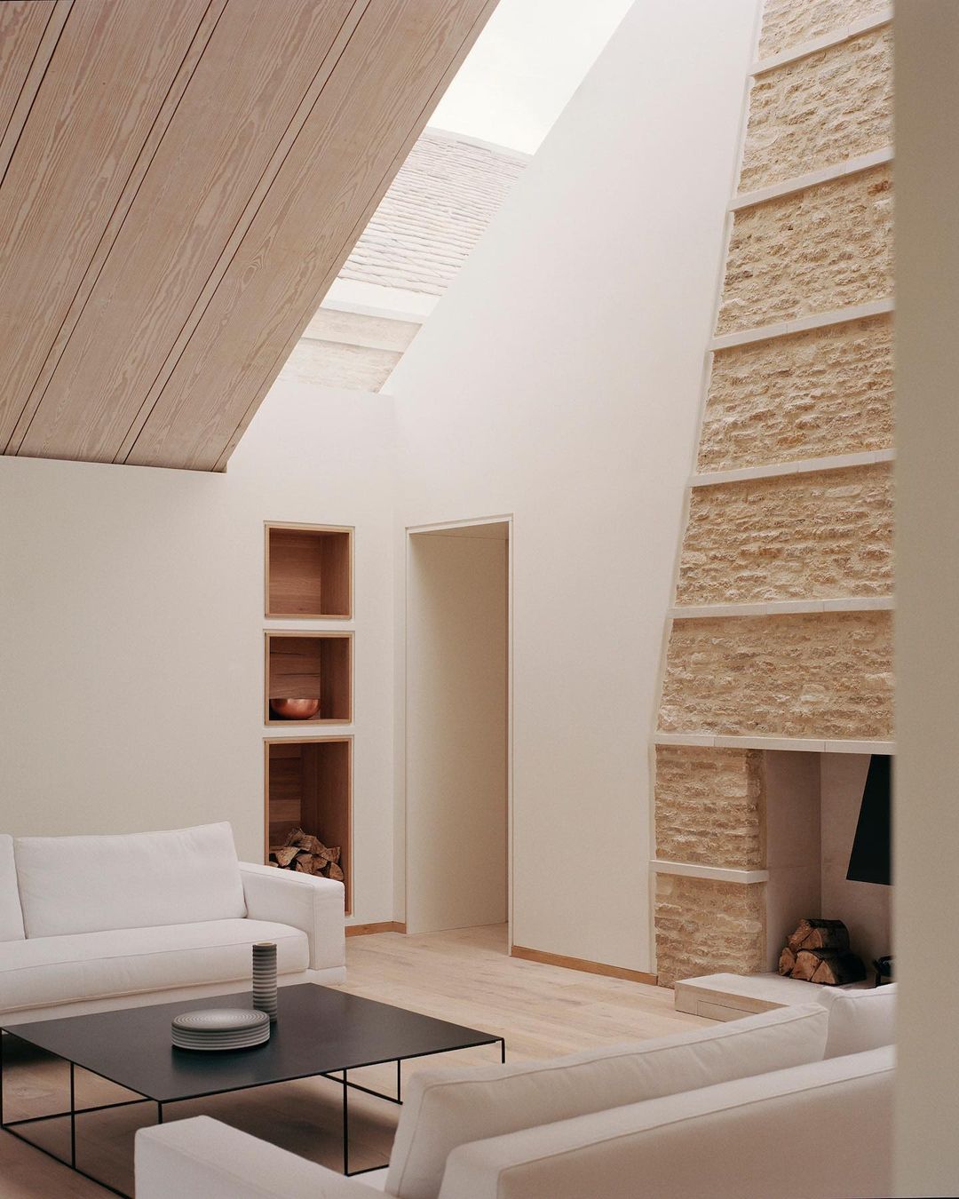 Contemporary living room interior with white sofas, black coffee table, stone fireplace, wood ceiling, bright open space with natural light.