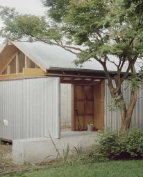 Corrugated metal shed with a pitched wooden roof framed by lush green trees and a garden under an overcast sky.