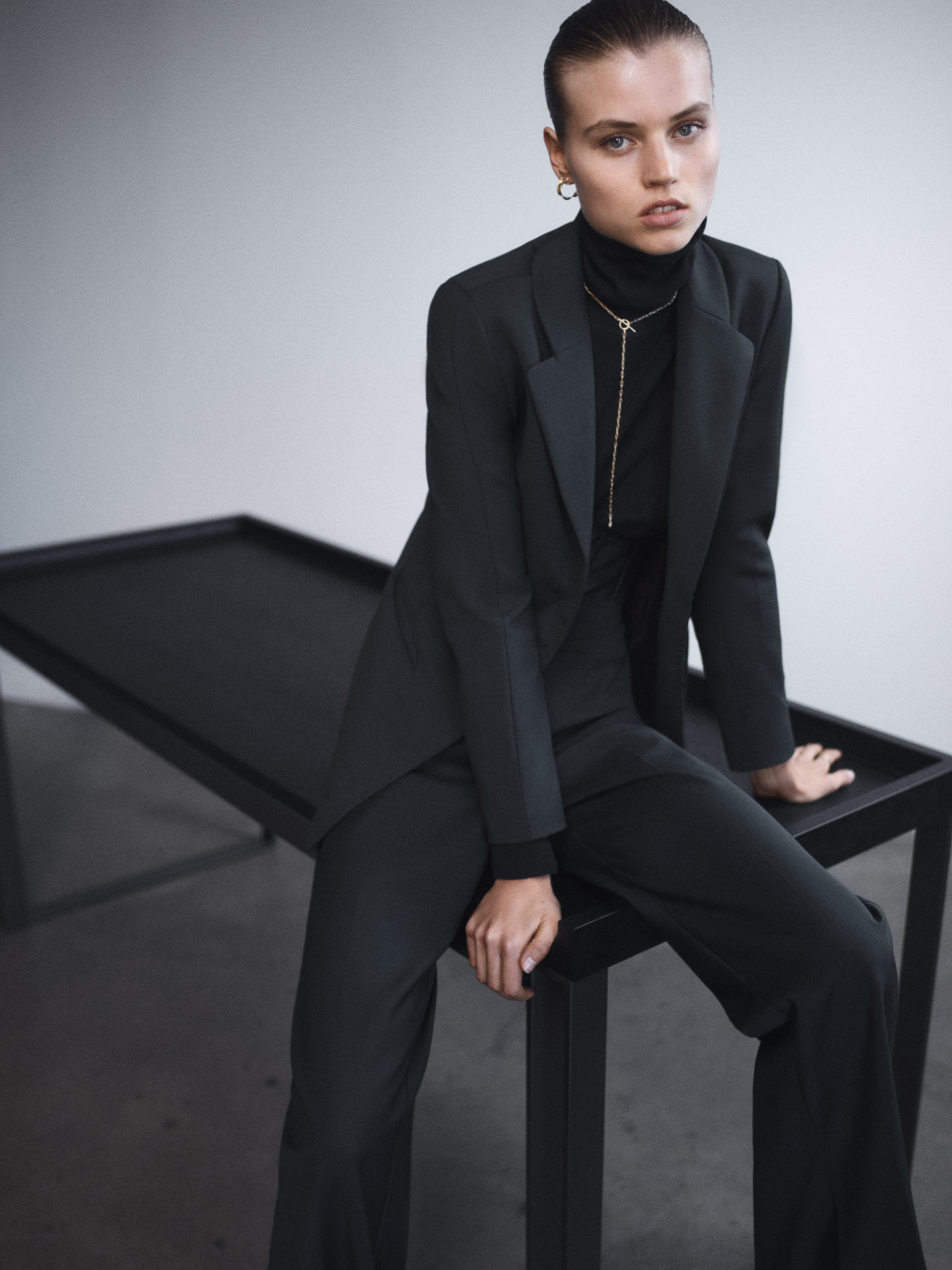 A stylish adult female model in a dark green blazer and wide-leg trousers sits on a black table in a minimalist studio setting.