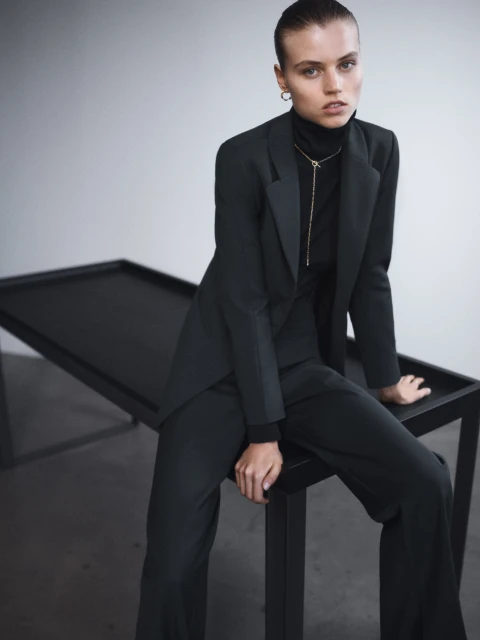 A stylish adult female model in a dark green blazer and wide-leg trousers sits on a black table in a minimalist studio setting.