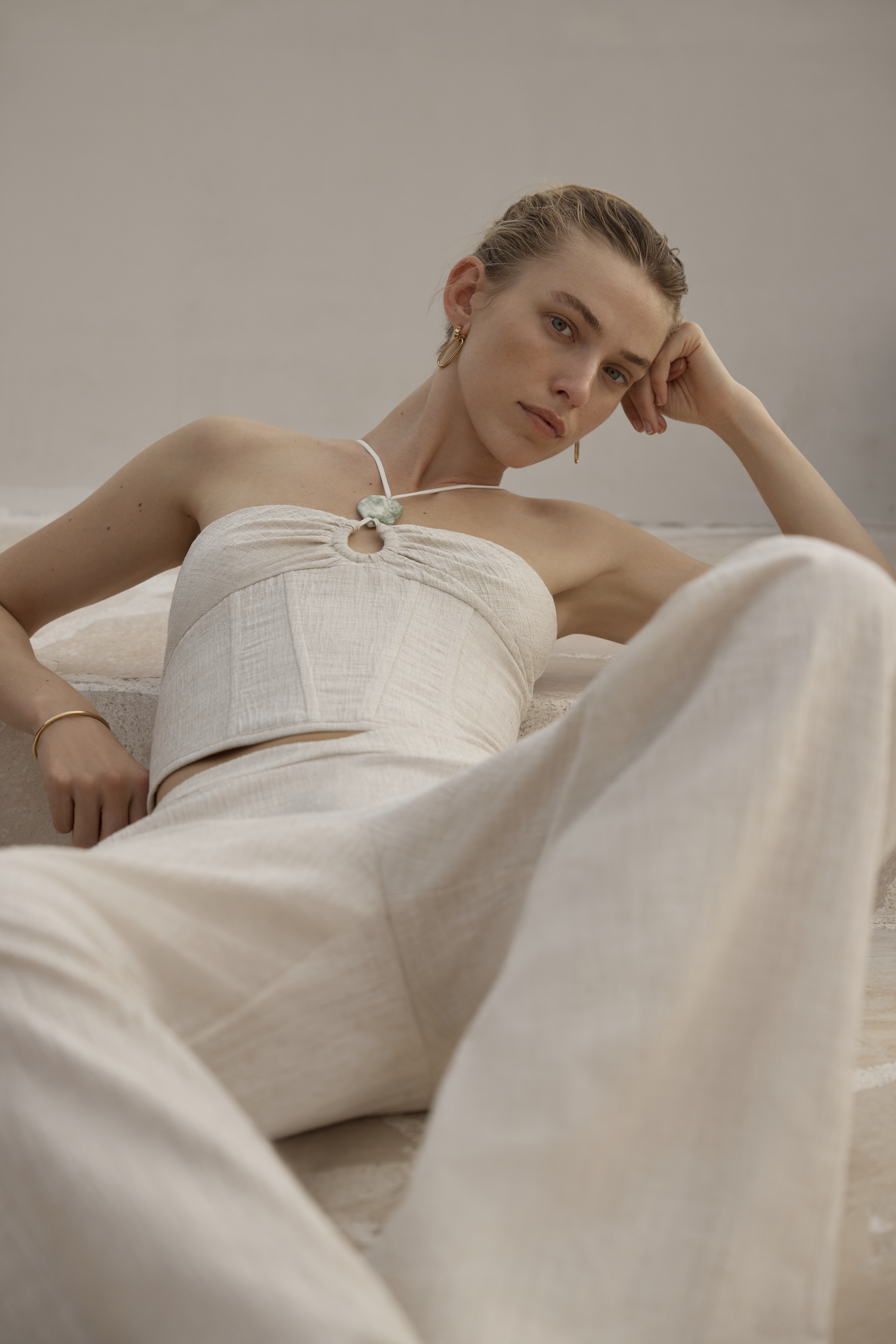 A woman in a light linen two-piece outfit reclines indoors. The outfit includes a strapless top and wide-legged pants in a neutral tone.