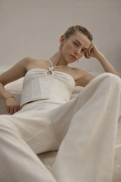 A woman in a light linen two-piece outfit reclines indoors. The outfit includes a strapless top and wide-legged pants in a neutral tone.