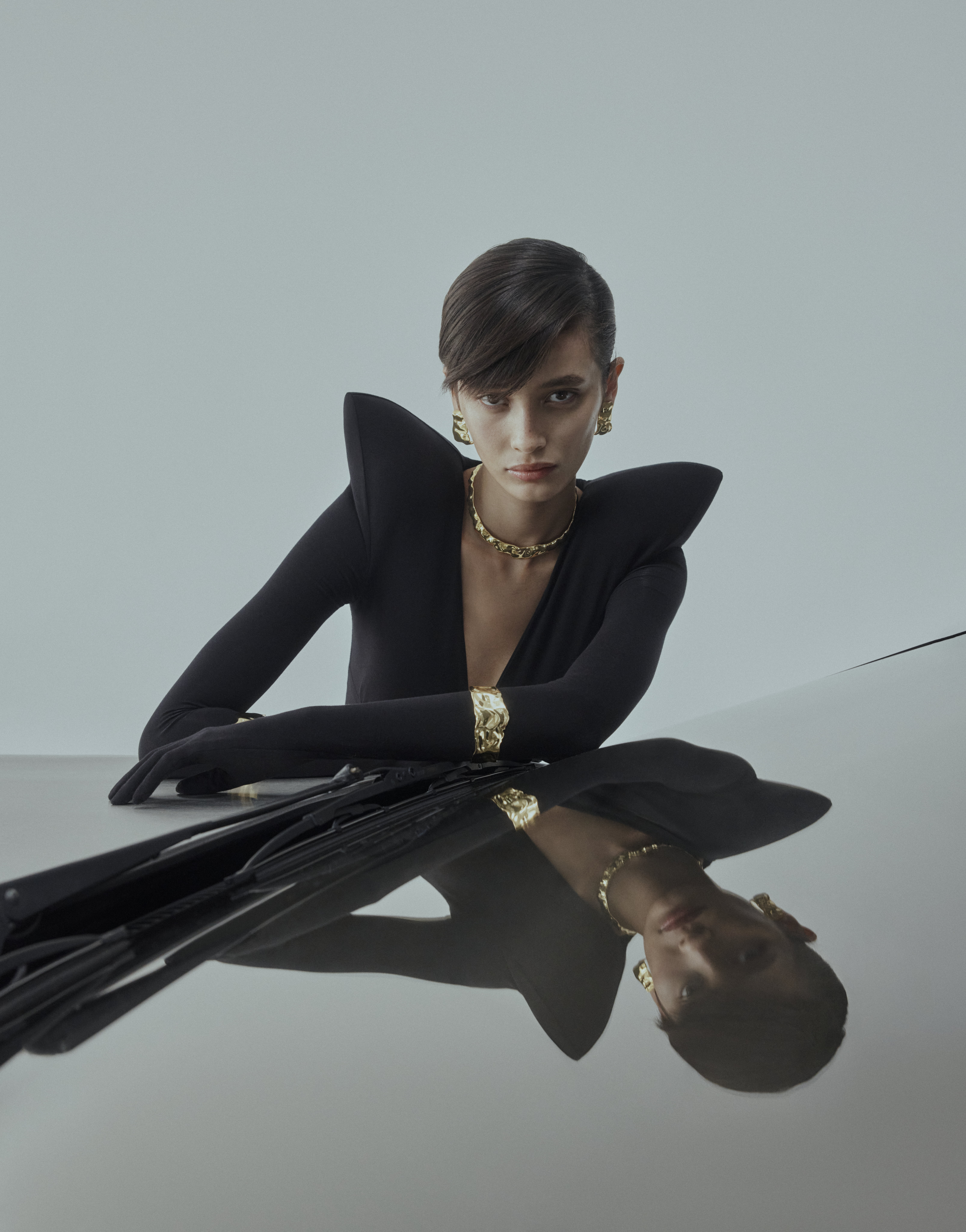 Brunette woman in long-sleeve black top with statement shoulders and gold jewelry poses with hands on reflective table.