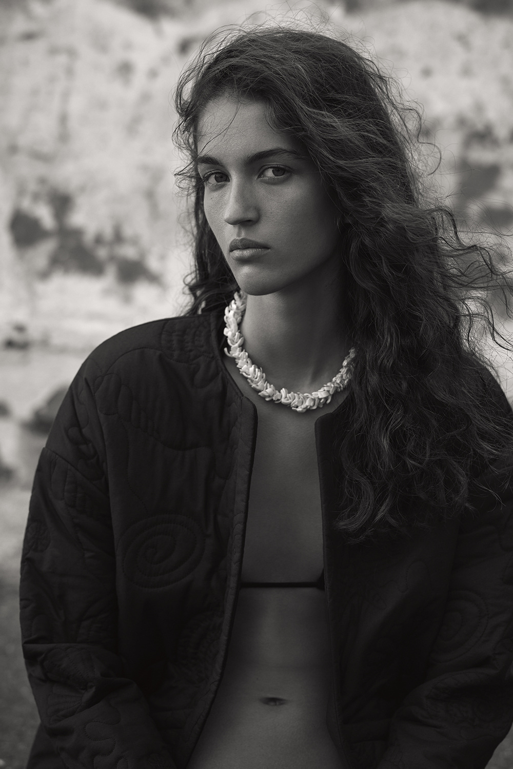Portrait of a young woman with long hair wearing a black jacket and a distinctive seashell necklace in an outdoor setting.