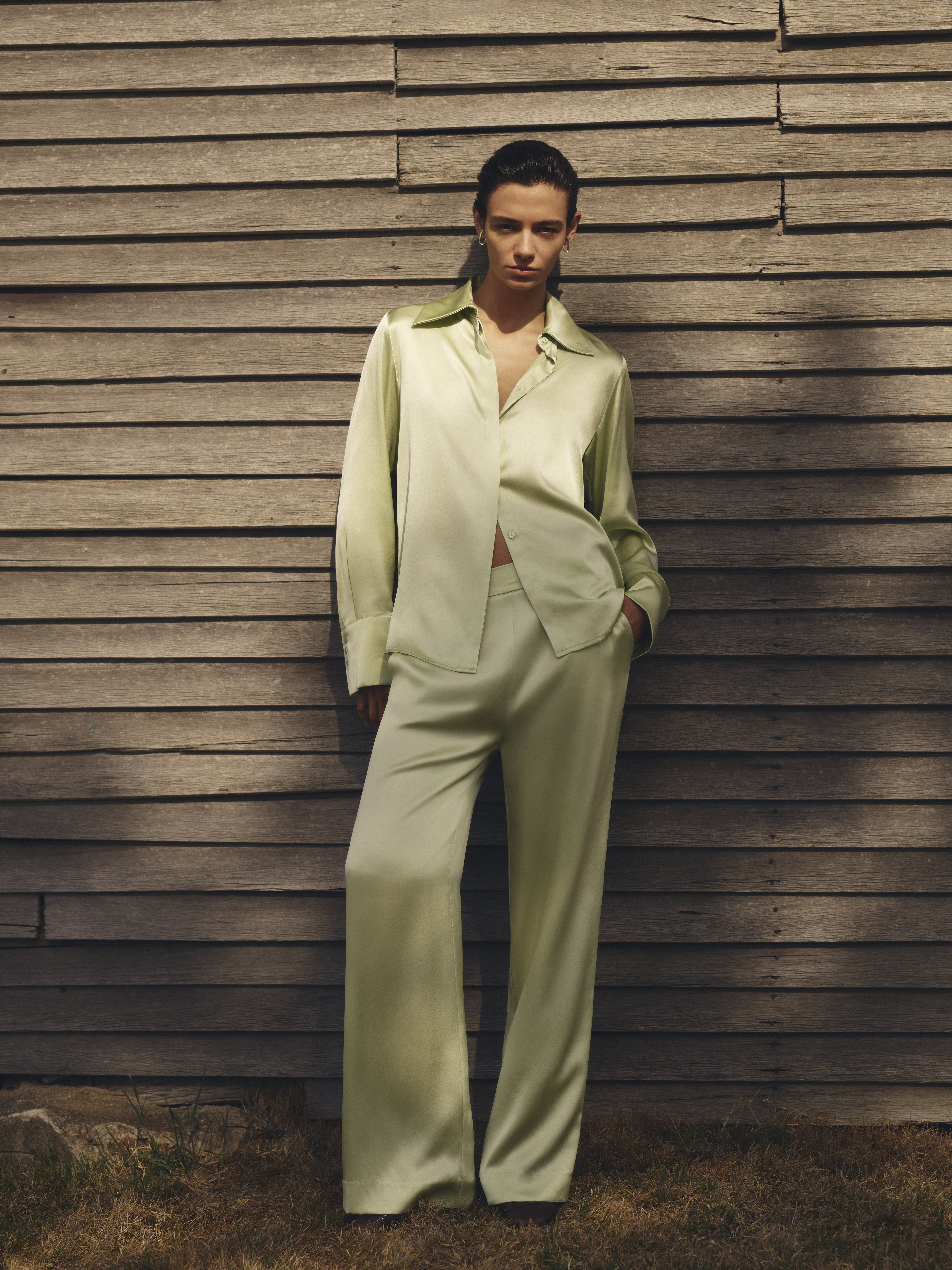 A young adult woman in a light green silky button-down shirt and pants stands against a wooden wall in an outdoor setting.