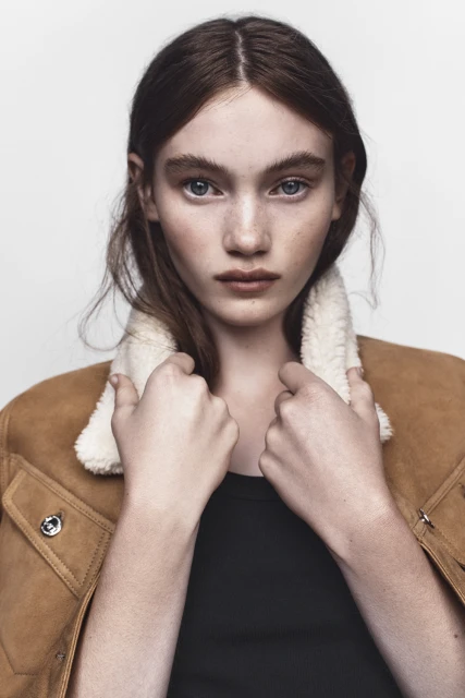 Close-up portrait of a young adult woman with auburn hair and blue eyes, wearing a tan coat with a white sherpa collar against a white background.