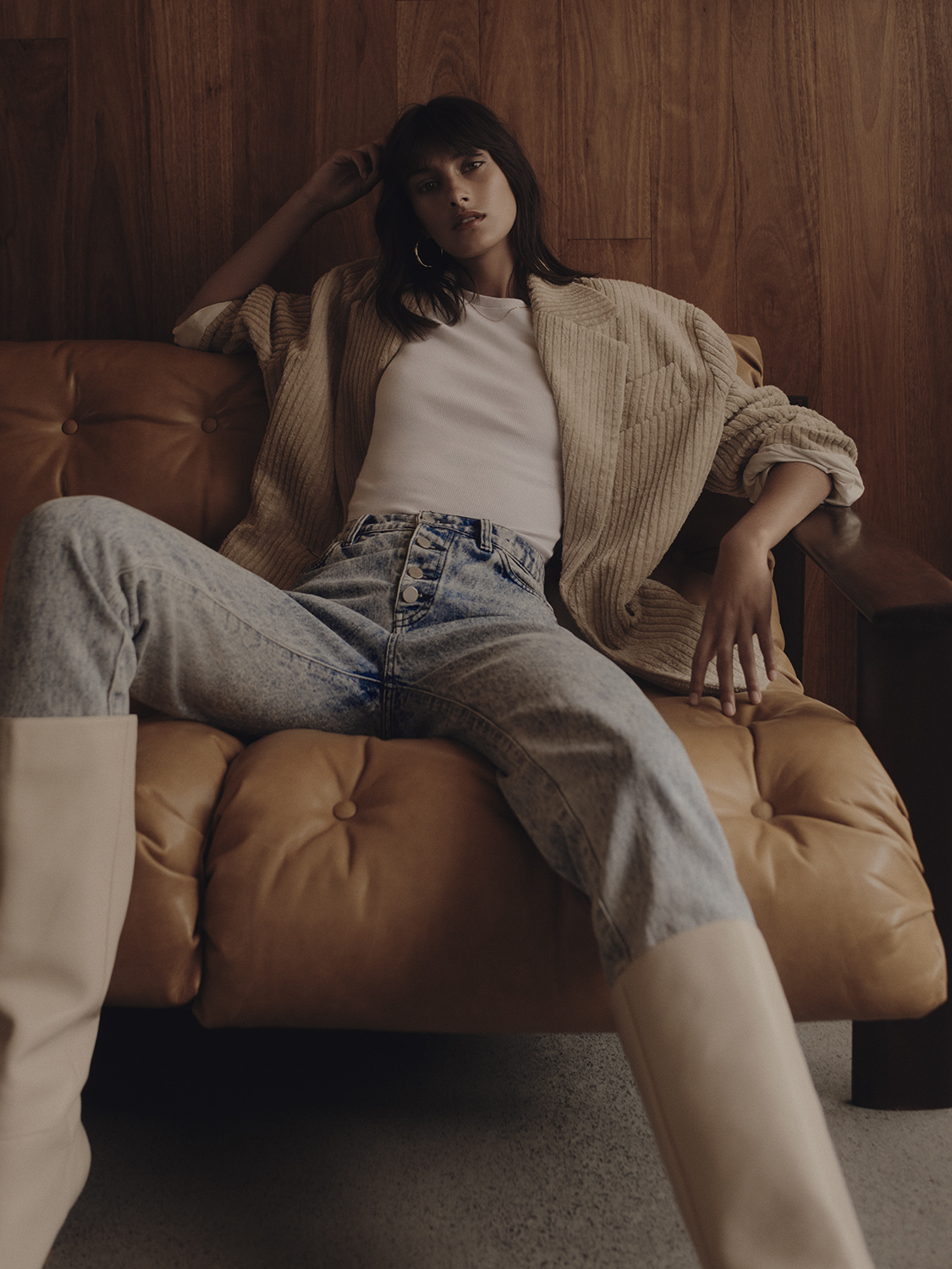 A woman lounges on a leather couch wearing a white t-shirt, acid wash jeans, a tan sweater, and tall boots in an indoor setting.