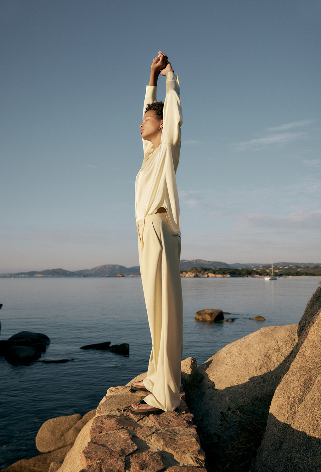 Woman in cream-colored blouse and wide-leg trousers with arms raised stands on rocks at the edge of a calm ocean under a clear sky.