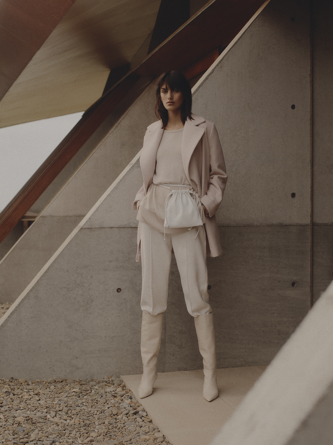 Fashionable woman poses in beige pantsuit, coat, and boots against a modern geometric concrete structure, in a neutral color palette.