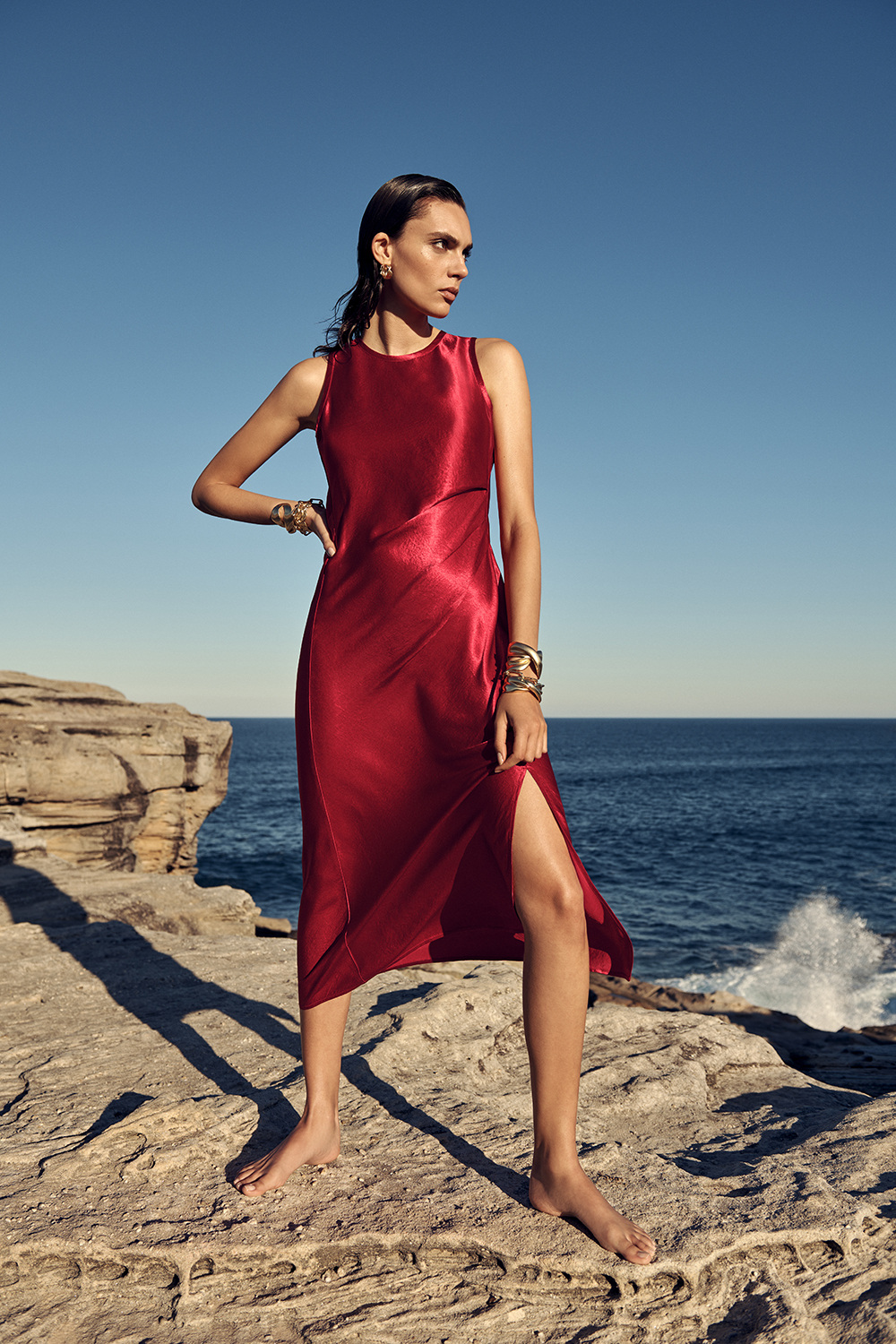 Woman in a red dress poses on a rocky shoreline against a blue sky and ocean backdrop, showcasing jewelry and a sleeveless, flowing design.