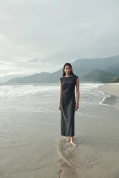 South Asian woman standing barefoot on a wet sandy beach wearing a dark sleeveless maxi dress with mountains in the background