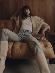 Woman in oversized beige blazer and denim jeans lounging on a leather sofa against wood paneling