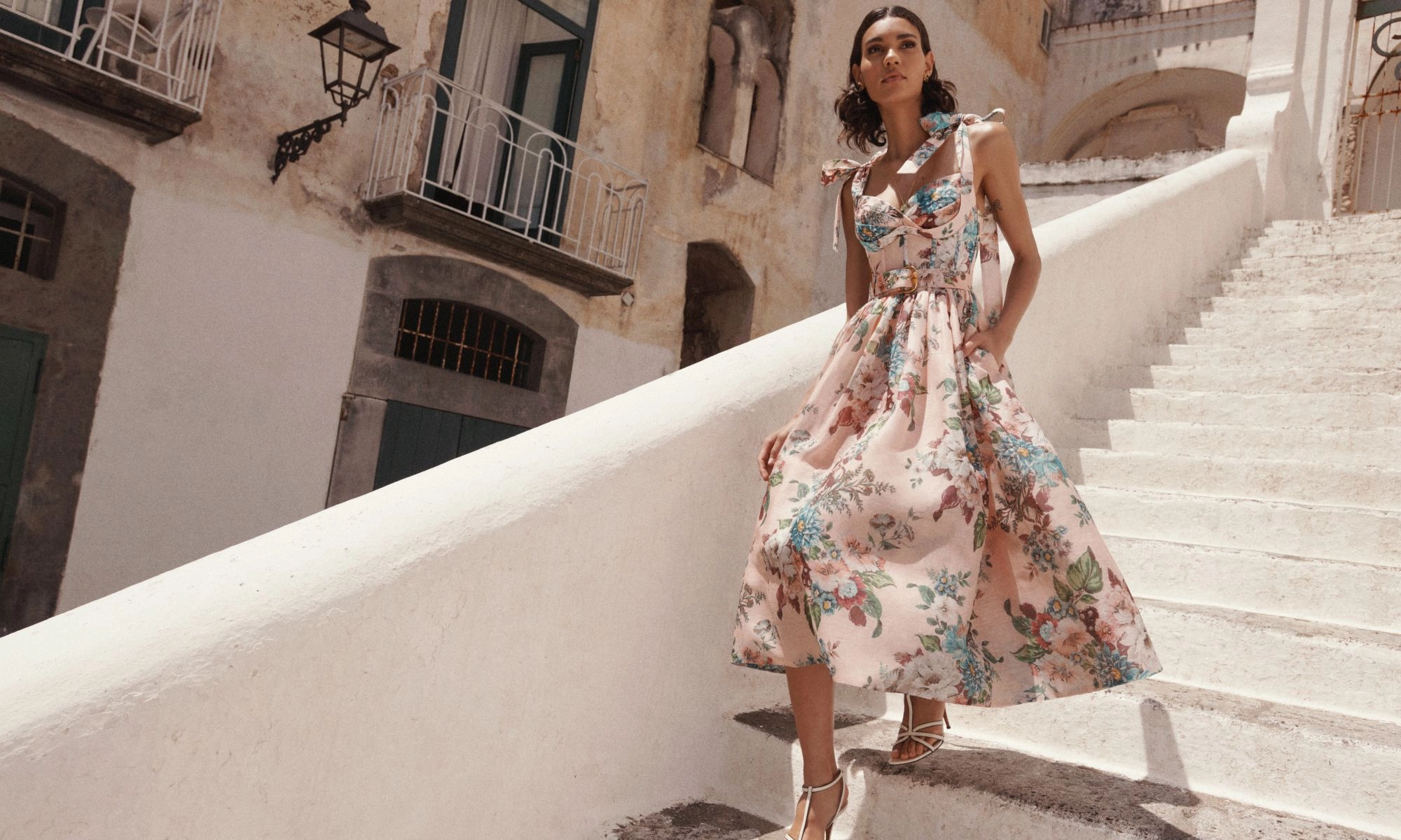 A woman wearing a floral dress walks down white stone steps in a sunlit outdoor setting with buildings in the background.