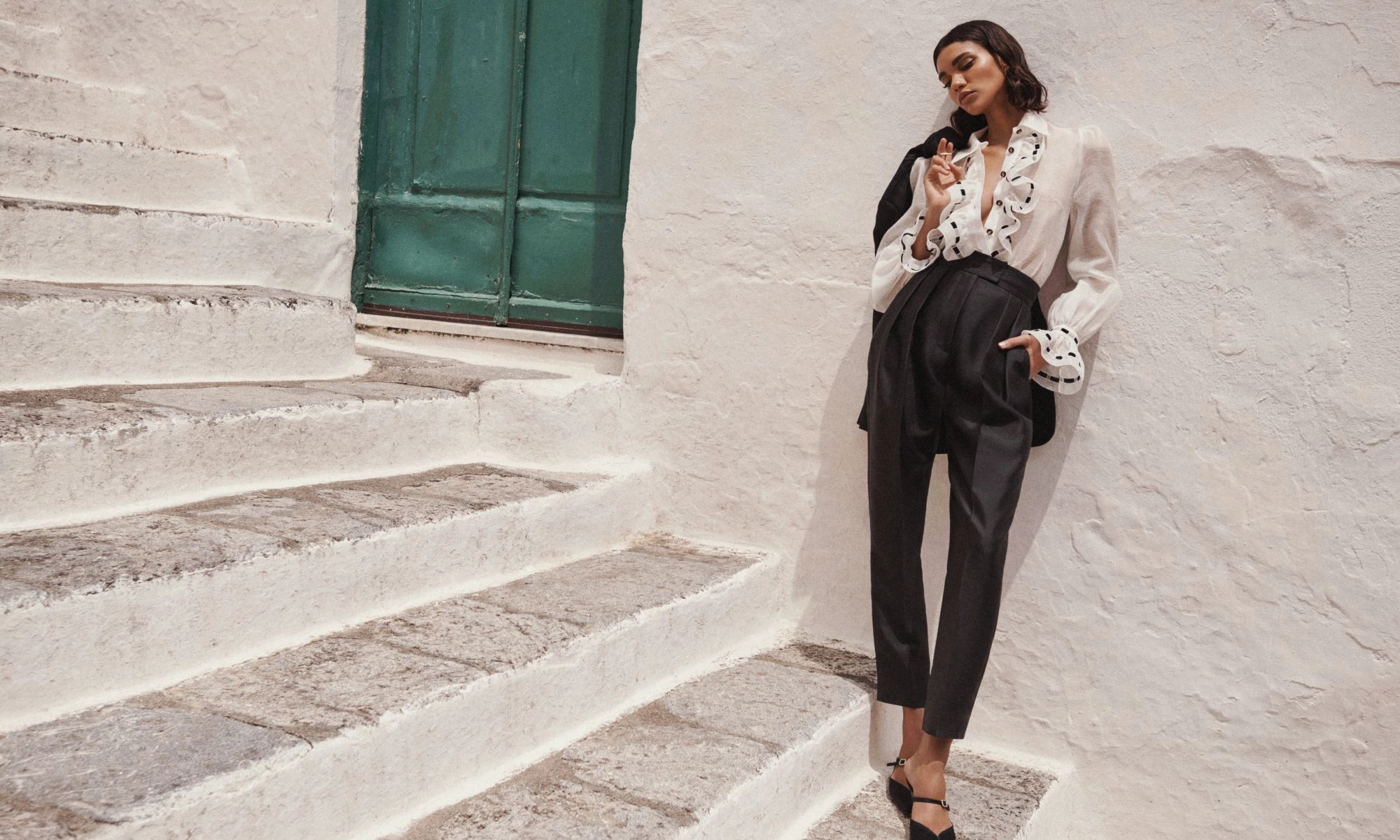 A fashion shot of a stylish woman posing on stone steps next to a white wall and a green door, showcasing a chic, high-fashion look.