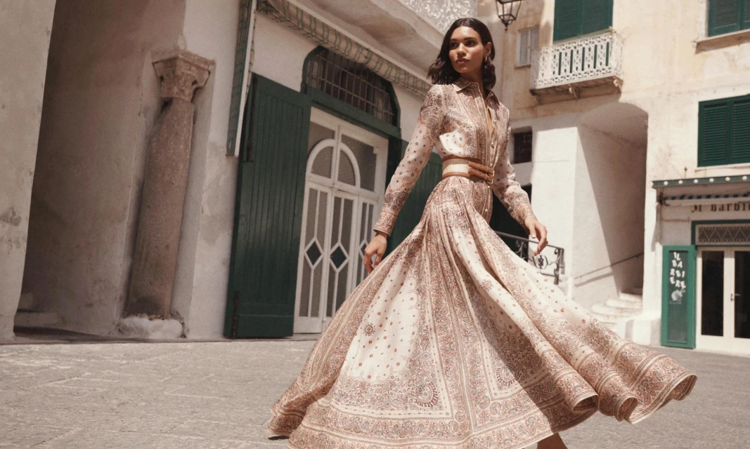 A brunette model wears a patterned maxi dress with a wide belt in an outdoor setting with rustic buildings and architecture in the background.