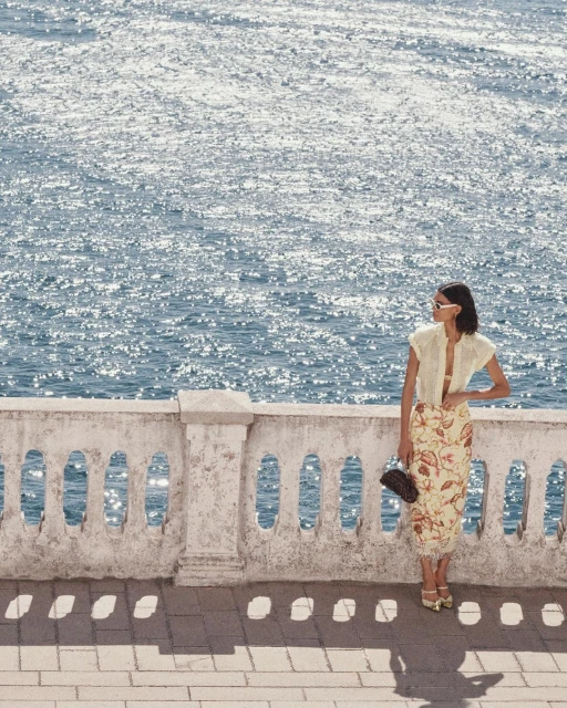 Woman in summer fashion stands by the sea on a sunlit stone terrace, wearing sunglasses and floral skirt with a clutch purse.