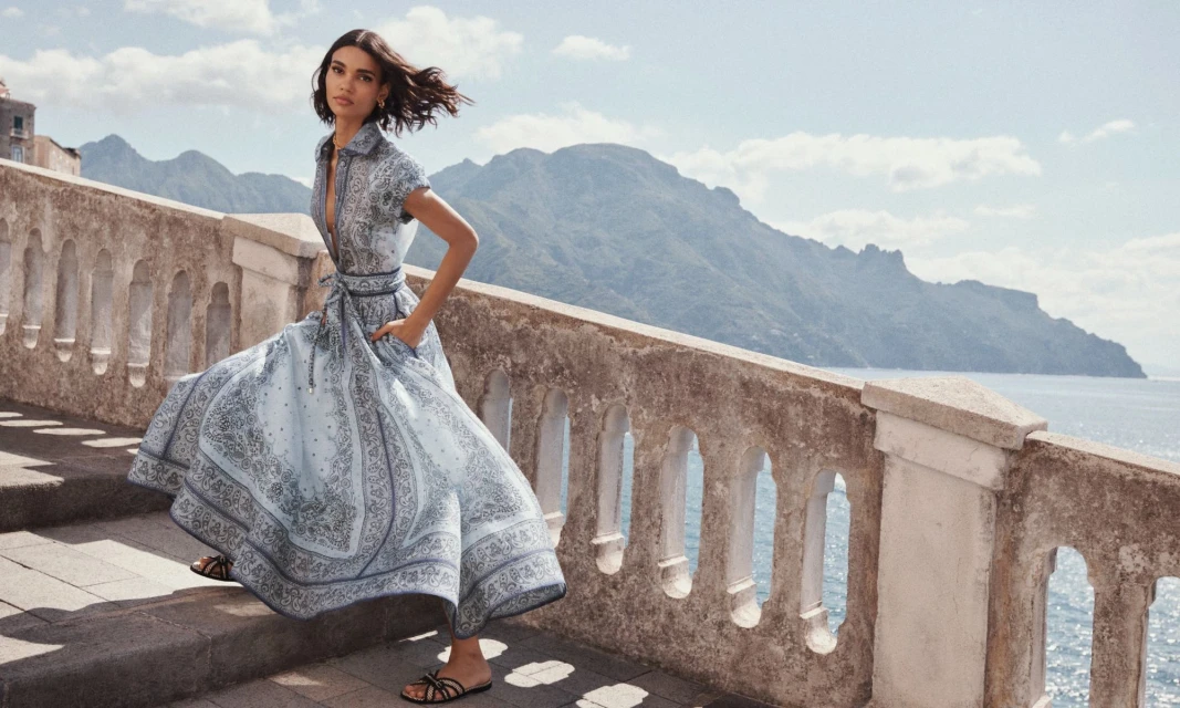 A woman in a patterned dress walks down stone steps against a backdrop of mountains and sea in a bright, outdoor fashion shot.