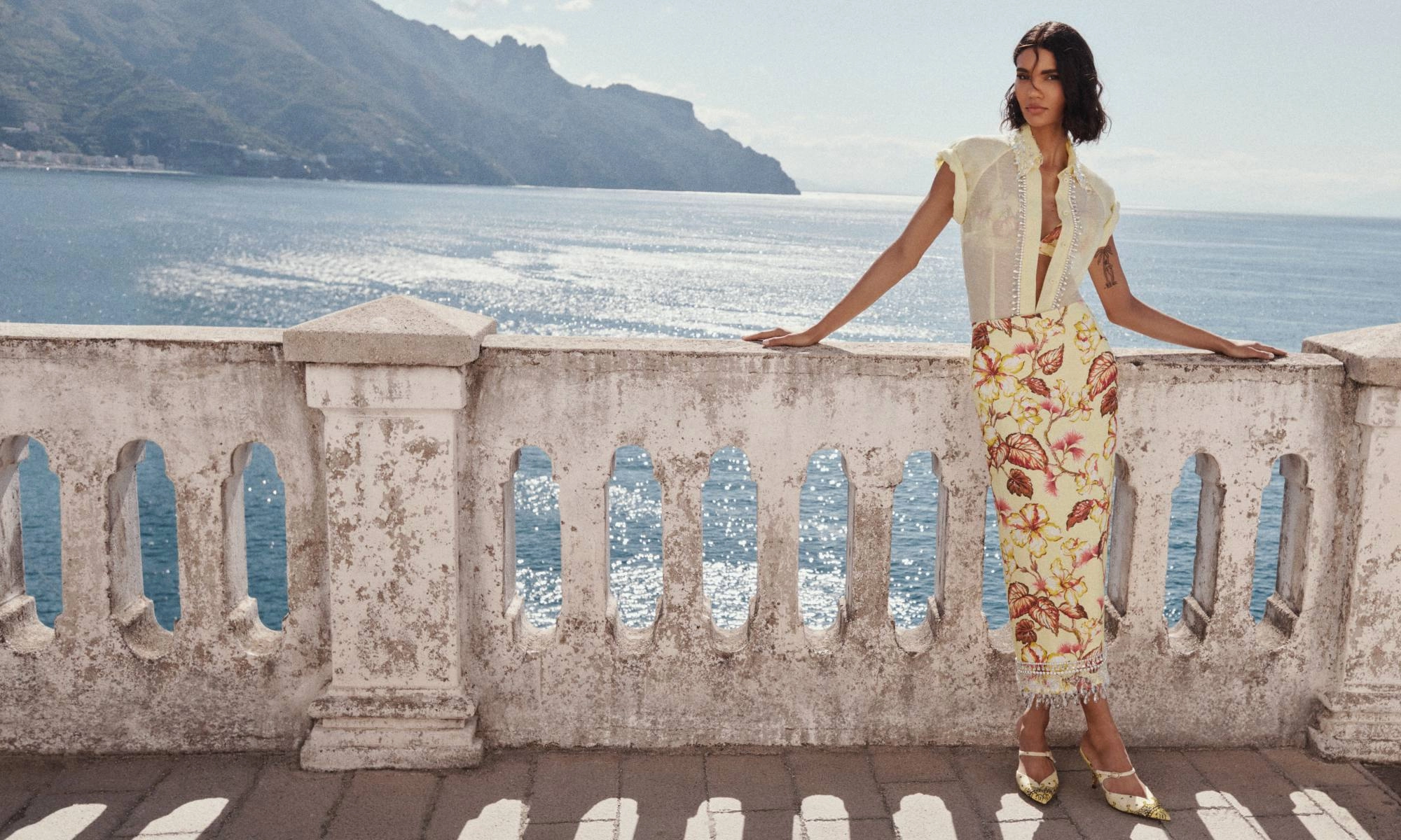 Brunette woman in sheer top and floral skirt poses on a balcony overlooking a blue sea with coastal mountains in the background.