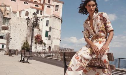 Model sitting on a park bench wearing a floral print dress holding a beaded handbag against a European street backdrop