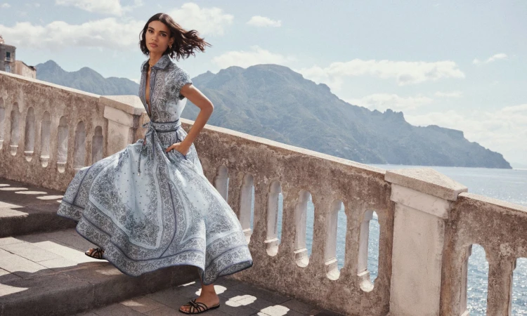Model in a patterned light blue belted midi dress standing on a stone balcony overlooking mountains and sea.